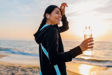 attractive slim woman doing sport exercises on morning sunrise beach in sports wear, thirsty drinking water in bottle, healthy lifestyle, listening to music on wireless earphones, feeling hot